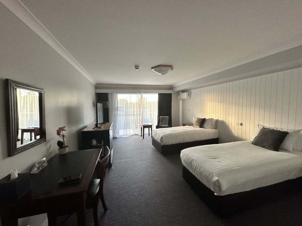 A spacious hotel room with two beds, a desk, and soft light coming through a window