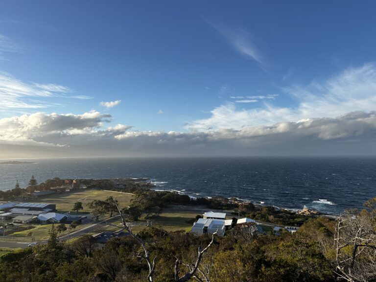 Coastal town seen from above, with low houses, open fields, and a deep blue ocean under a wide sky with scattered clouds.